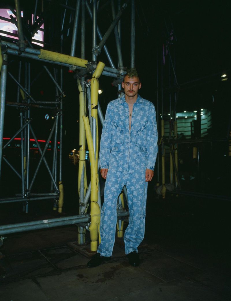 A young adult male stands outdoors at night in a printed blue jumpsuit with metal scaffolding and lights in the urban background.