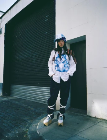 Woman in streetwear fashion poses in front of a closed roll-up door, wearing a bucket hat, graphic tee, and platform shoes with ribbons.