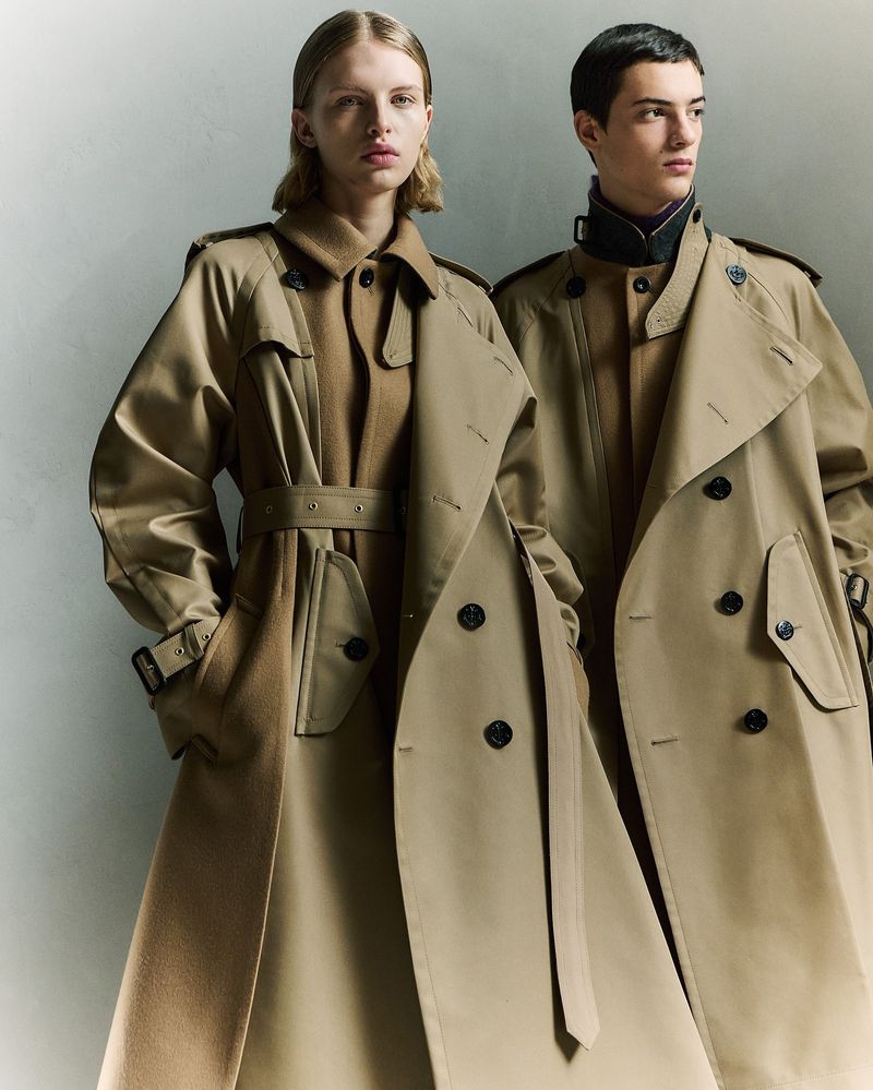 Two people, a woman and a man, standing in front of a neutral backdrop, both wearing long tan trench coats, fashion shot, studio lighting.