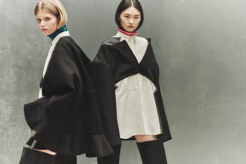 Two women in avant-garde fashion stand against a textured grey background, showcasing oversized black and white layered outfits and neutral makeup.