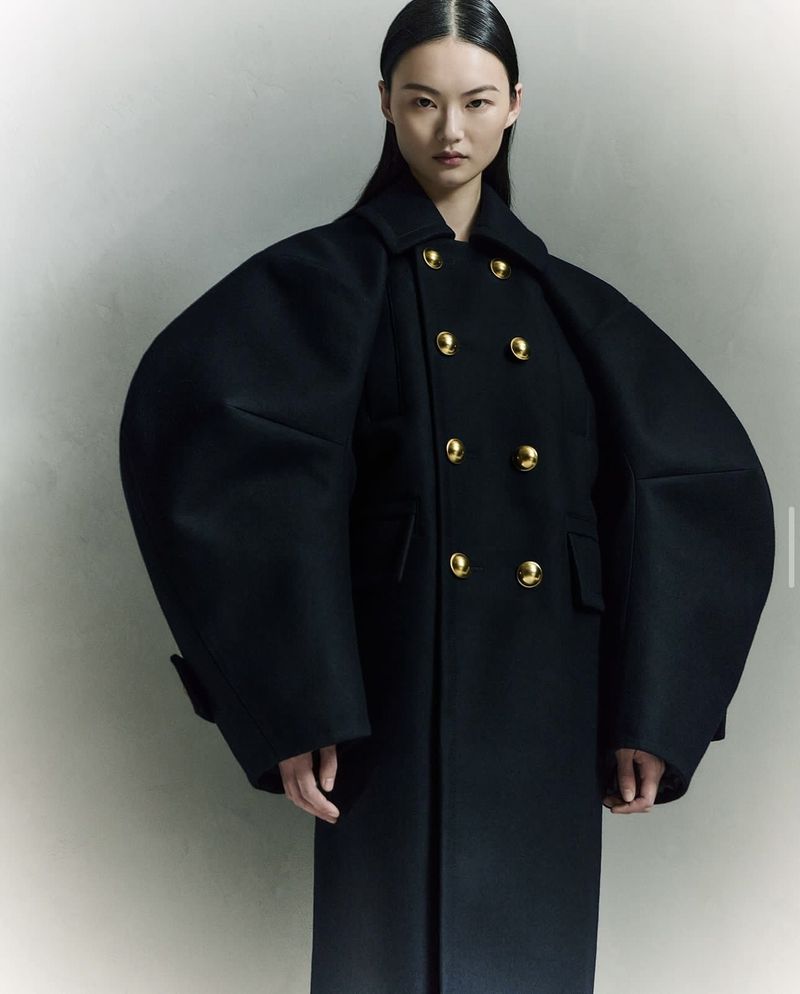 An Asian woman poses indoors wearing a long black double-breasted wool coat with gold buttons, styled with sleek, straight black hair.