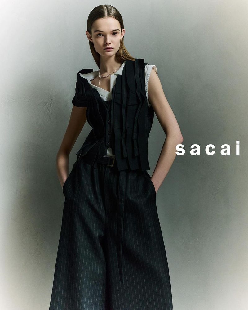A woman poses in a sleeveless black top and pinstripe pants with her hands in her pockets against a gray studio backdrop.