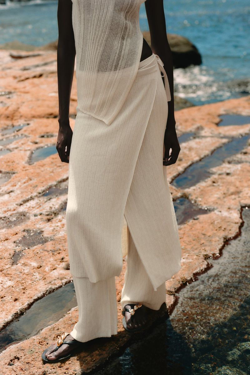 A woman models off-white knitwear on a rocky shore, featuring a sleeveless top, wrap-style pants, and black sandals in a fashion editorial.