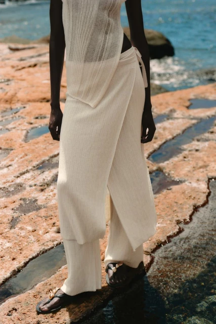 A woman models off-white knitwear on a rocky shore, featuring a sleeveless top, wrap-style pants, and black sandals in a fashion editorial.