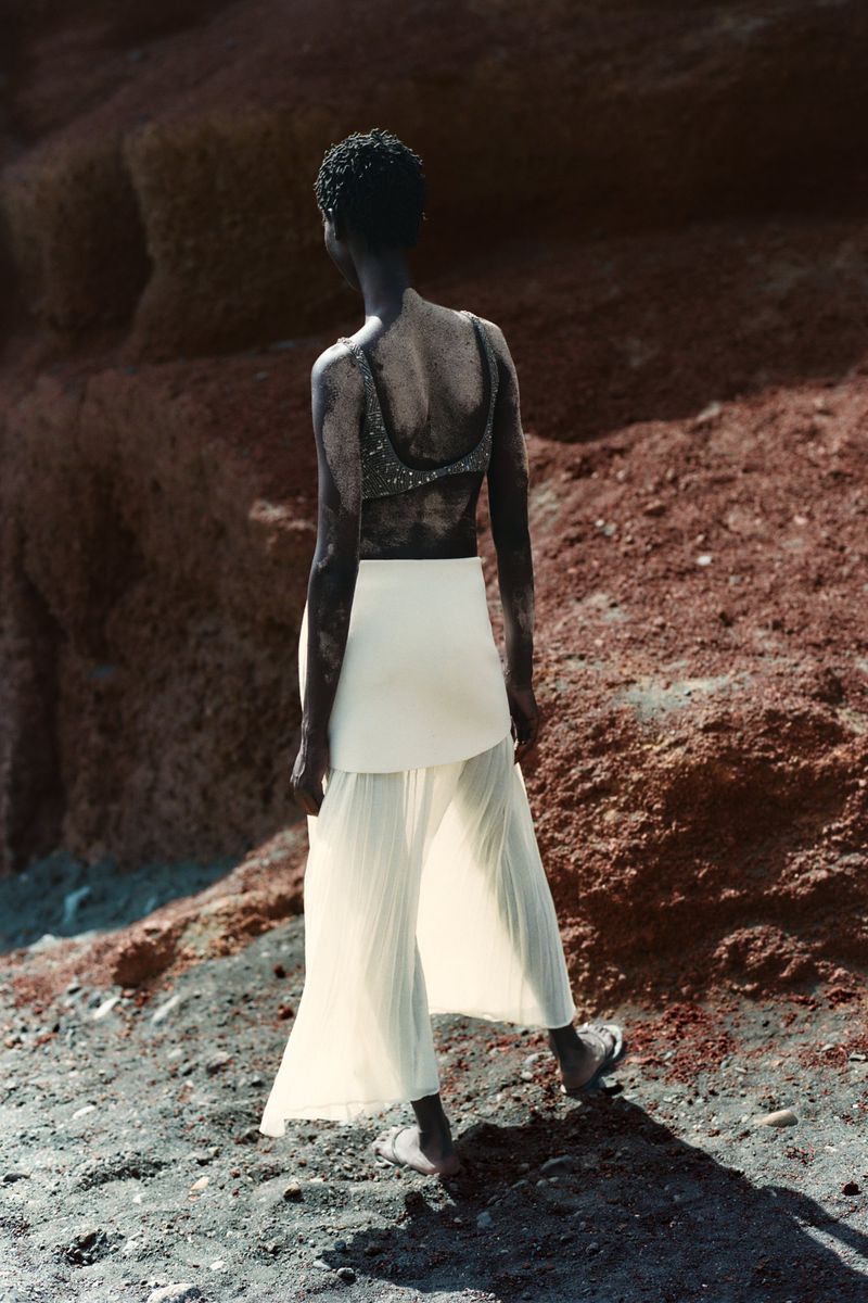 An adult female model stands on a rocky surface, wearing a shimmering top and loose, white trousers, viewed from the back in outdoor light.