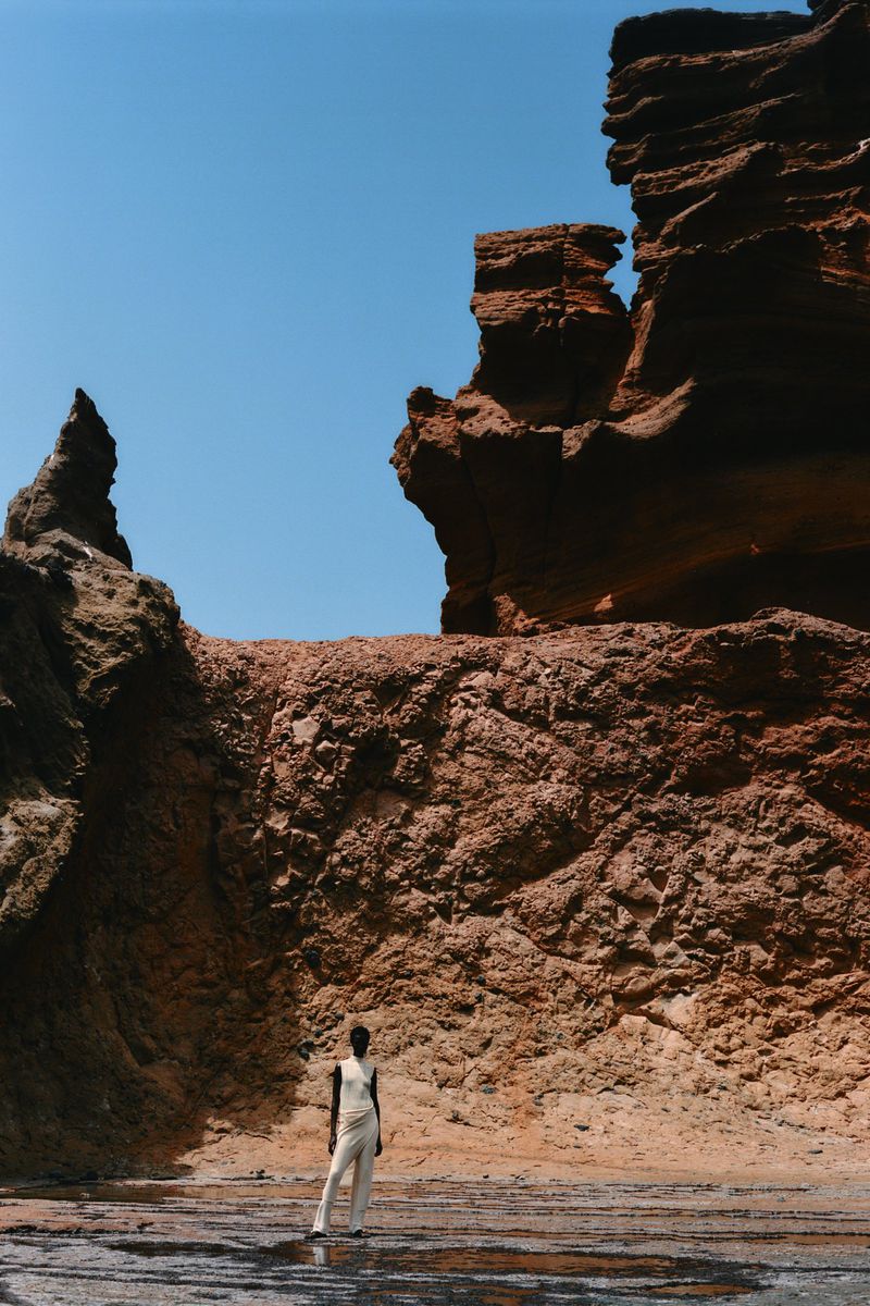 Woman in neutral jumpsuit standing on rocky surface with textured rock formations, blue sky, on sunny day in outdoor environment.