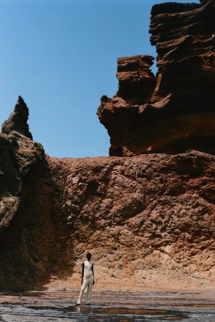 Woman in neutral jumpsuit standing on rocky surface with textured rock formations, blue sky, on sunny day in outdoor environment.