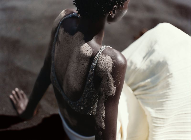 Woman with dark skin poses on a sandy beach. Her back and shoulders are covered in sand. She wears a beaded top and a white skirt.