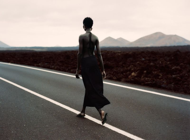 Woman in long black dress walks barefoot on an empty road with distant mountains, displaying a fashion and travel aesthetic.