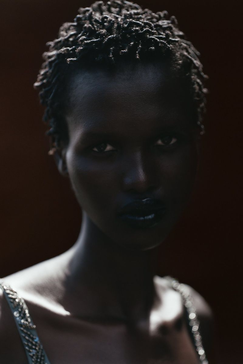 Close-up studio shot of a dark-skinned woman with short curly black hair and dark makeup, wearing a beaded top against a dark background.
