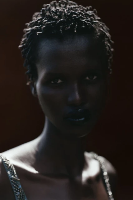 Close-up studio shot of a dark-skinned woman with short curly black hair and dark makeup, wearing a beaded top against a dark background.