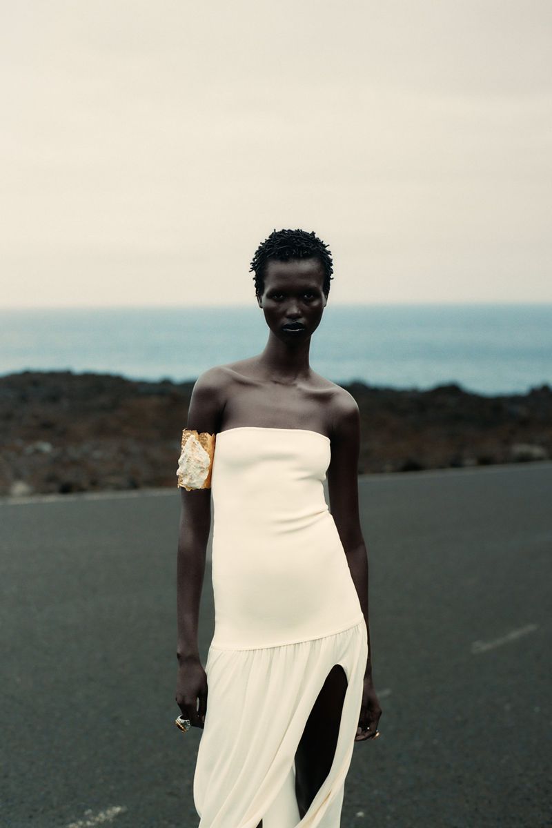 Woman in a cream strapless dress and arm cuff stands on a black surface, with a blurred background of ocean and sky.