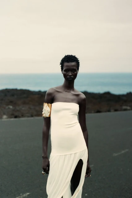 Woman in a cream strapless dress and arm cuff stands on a black surface, with a blurred background of ocean and sky.
