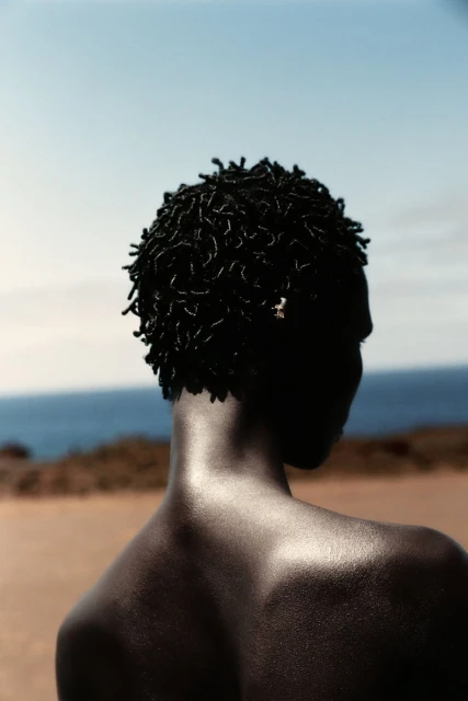 Back view of an African American adult with short dreadlocks outdoors. The scene features a blurred coastline and clear blue sky.