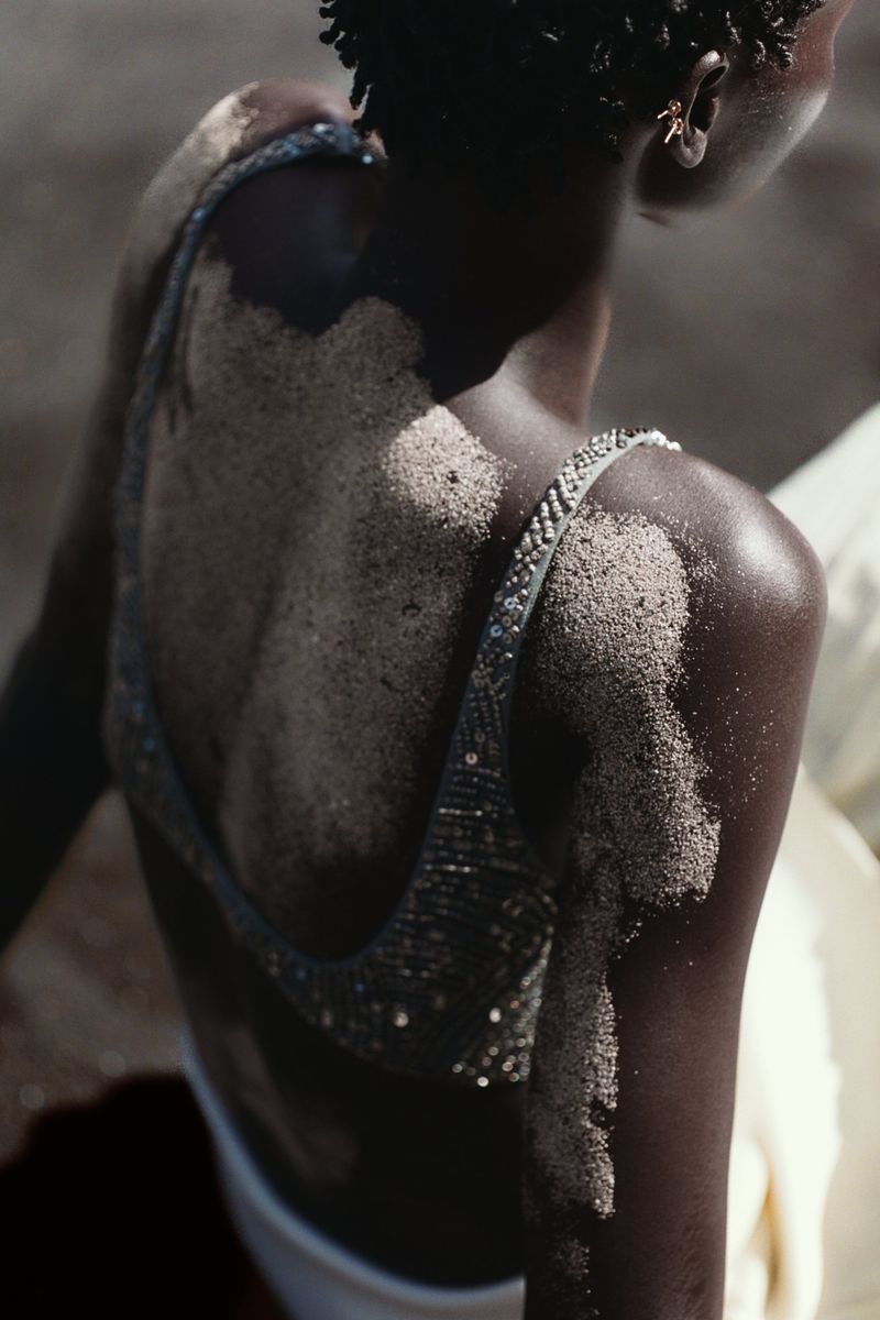 An adult female model with dark skin poses in a sand-covered top and white bottoms, viewed from the back, against a blurred outdoor background.