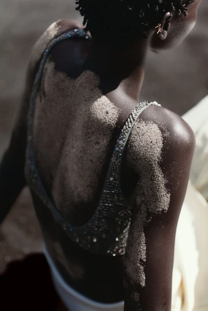 An adult female model with dark skin poses in a sand-covered top and white bottoms, viewed from the back, against a blurred outdoor background.