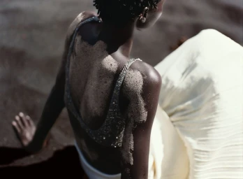 Model Amar Akway seated on dark sand with sand on her bare back, wearing an embellished top and off-white pleated skirt.