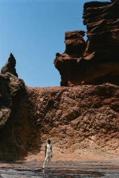 Model Amar Akway wearing an off-white ensemble standing before towering red rock cliffs under a clear blue sky.