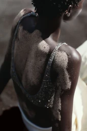 Model Amar Akway seen from behind with textured sand across her bare back and shoulder, wearing a beaded sequin top.