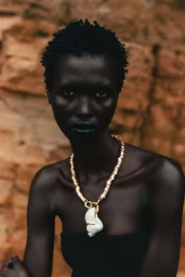 Model Amar Akway wearing a gold shell necklace and dark top against a natural textured rock wall background
