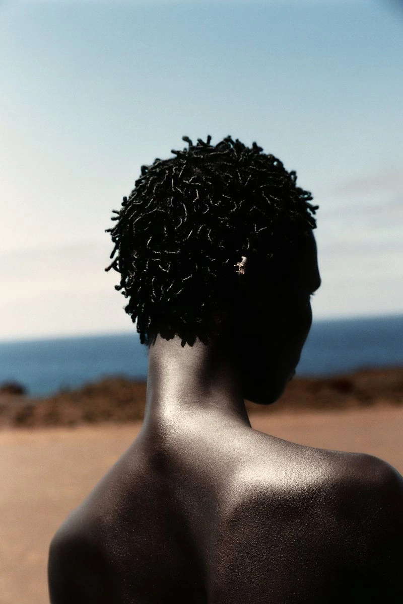 Model Amar Akway seen from behind with short textured curls against a sunlit coastal backdrop.