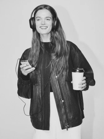 Woman with headphones smiles, holding a coffee cup and smartphone in a black and white portrait-style image, against a light background.