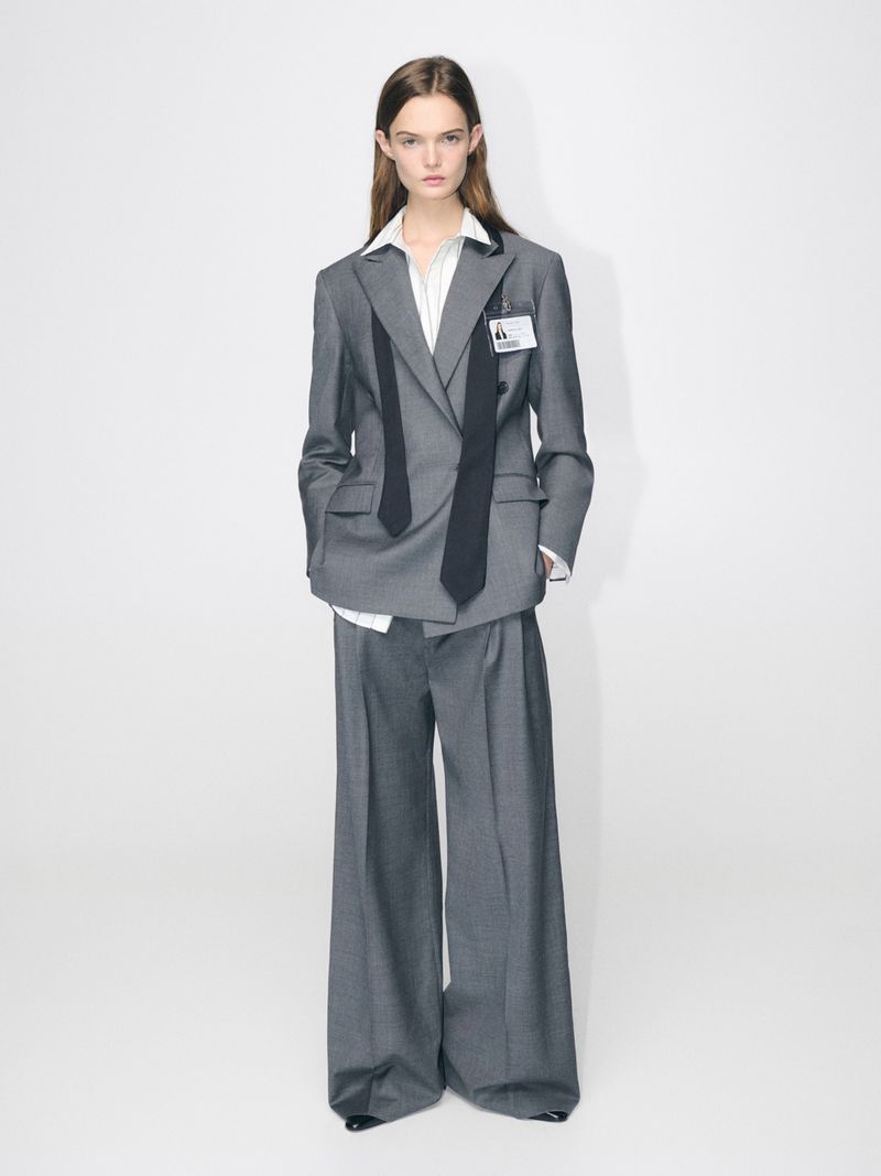 Full-length studio shot of a model wearing a grey pantsuit with a black tie detail, and a white background, showcasing a modern fashion look.