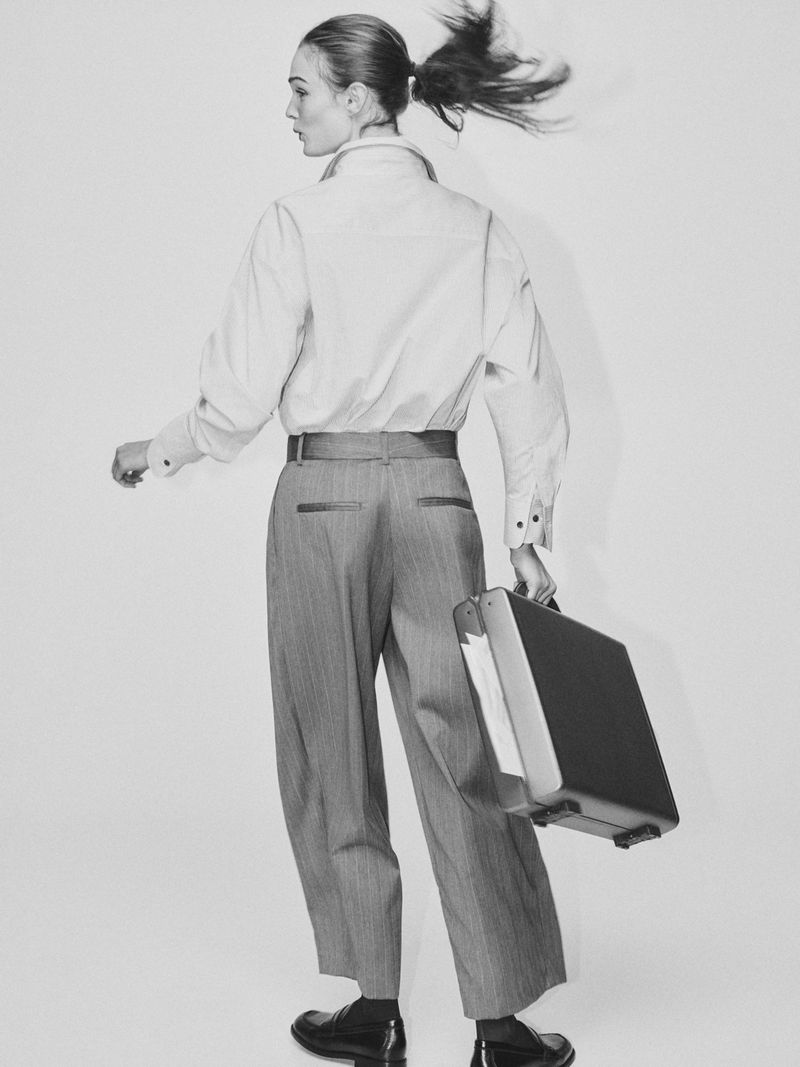 Stylish woman in button-down shirt and wide-leg pants carries a briefcase and glances to the left in a monochrome studio portrait.