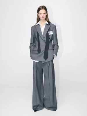 Model Lulu Tenney posing in an oversized grey trouser suit with a draped black necktie against a plain white studio background.