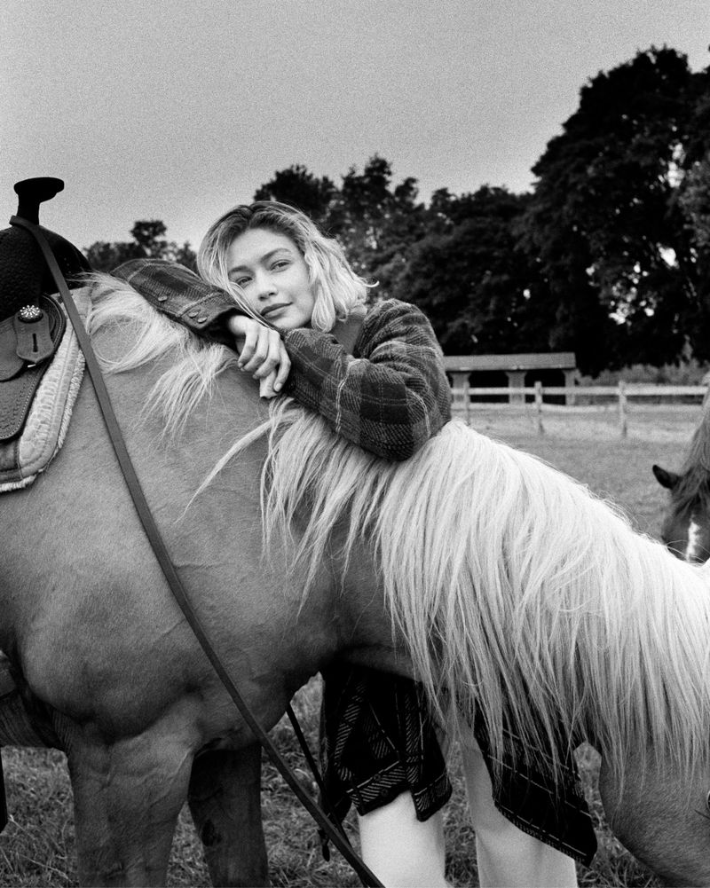 Black and white photograph of an adult woman wearing a plaid jacket lying across a horse in an open field in a rural setting.
