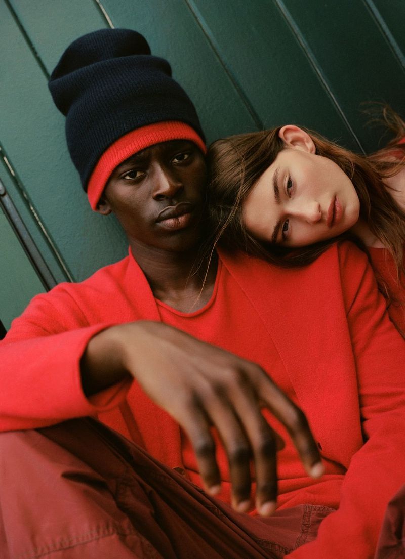 A dark-skinned man in a beanie and a fair-skinned woman pose closely in red shirts against a green background in a fashion shot.