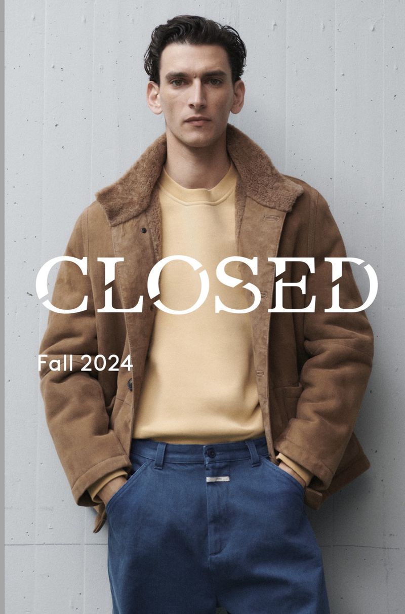 A fair-skinned male model poses in a brown jacket, yellow sweater, and blue jeans against a gray wall. Fashion advertisement for Fall 2024.