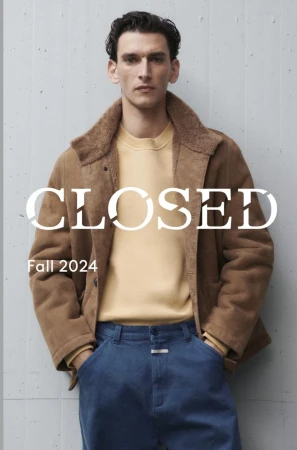 A fair-skinned male model poses in a brown jacket, yellow sweater, and blue jeans against a gray wall. Fashion advertisement for Fall 2024.