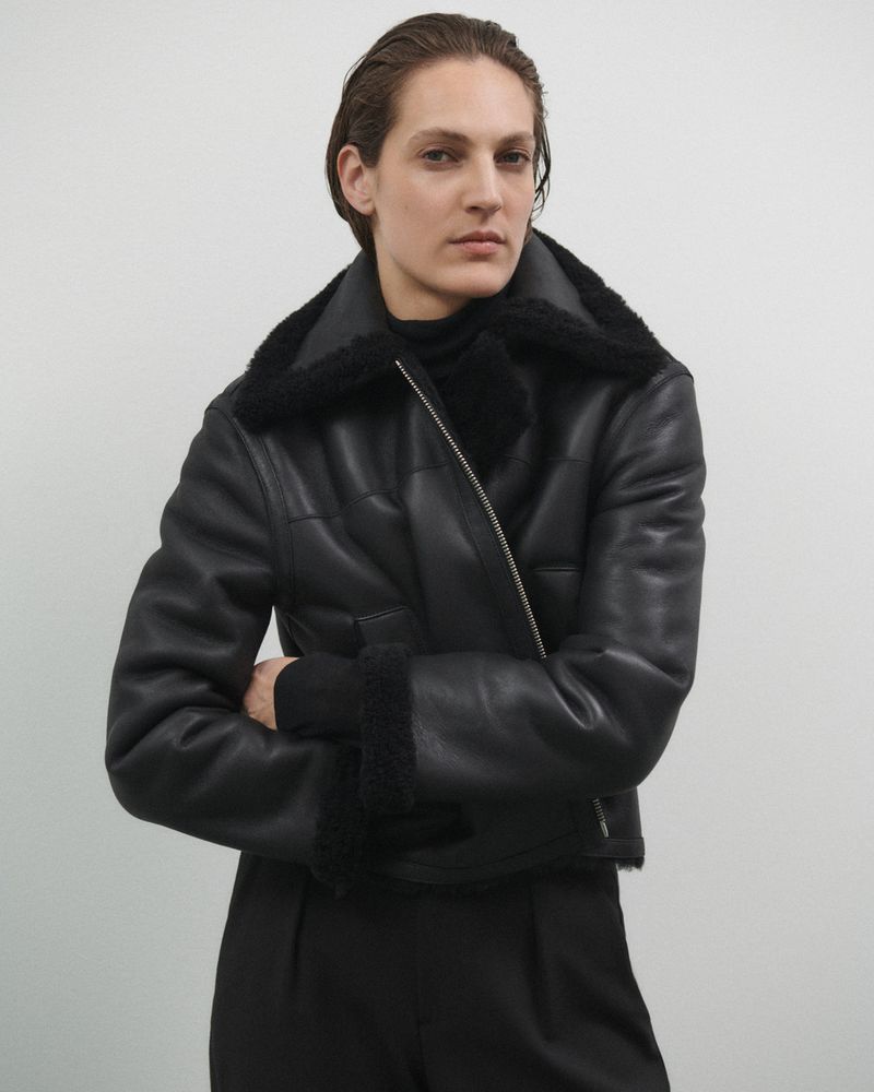 Woman poses indoors wearing a black shearling jacket and trousers. The neutral background places emphasis on the fashion garment.