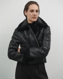 Model wearing a black leather jacket with a black shearling collar against a neutral grey studio background