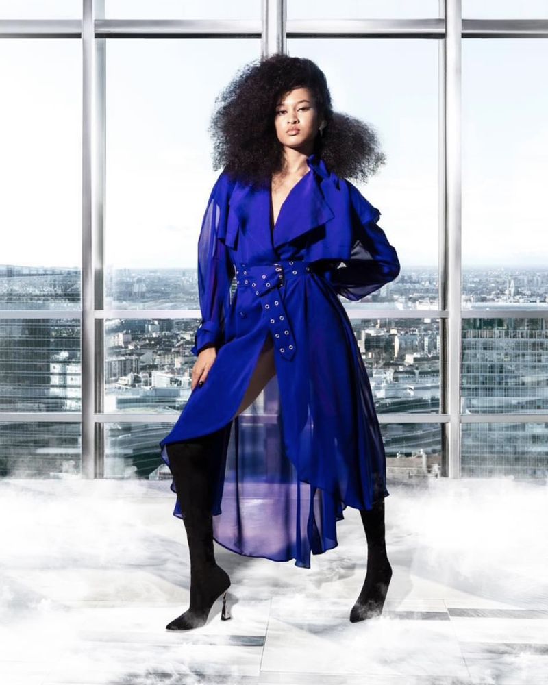 Adult woman in blue trench coat, thigh-high boots poses in a light-filled indoor space with a cityscape view beyond large windows.
