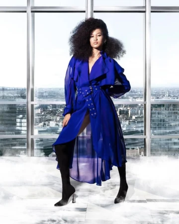 Adult woman in blue trench coat, thigh-high boots poses in a light-filled indoor space with a cityscape view beyond large windows.