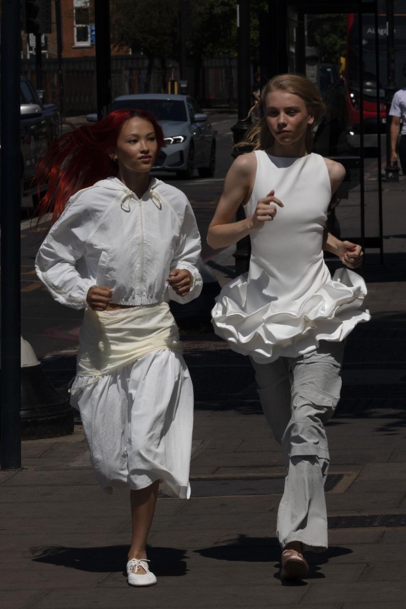 Two women in white outfits run on a city sidewalk on a sunny day, captured in a medium shot showcasing fashion and urban lifestyle.