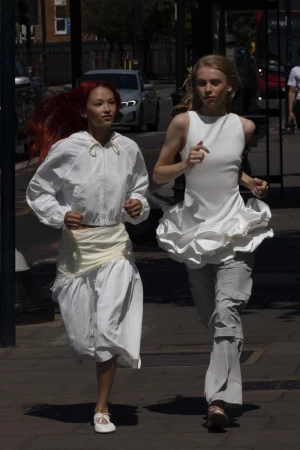 Two women in white outfits run on a city sidewalk on a sunny day, captured in a medium shot showcasing fashion and urban lifestyle.