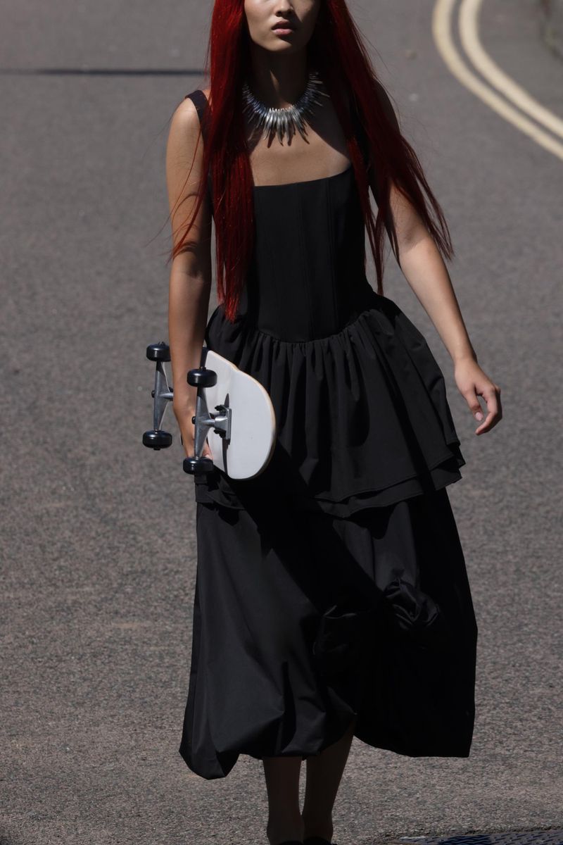 A female model with red hair walks on an asphalt road in a black dress, holding a skateboard, creating a striking fashion image.