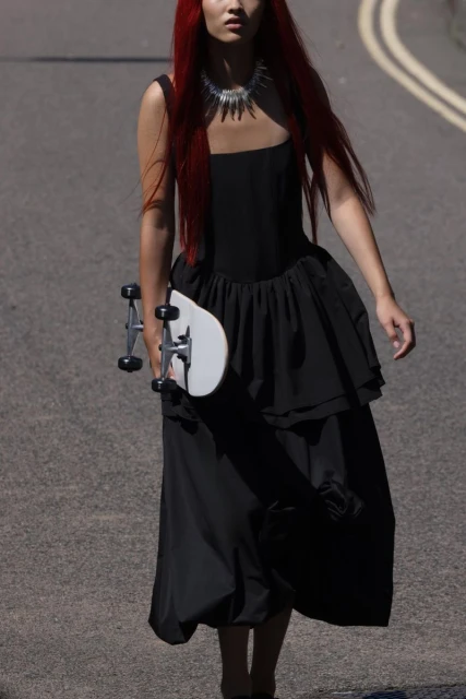 A female model with red hair walks on an asphalt road in a black dress, holding a skateboard, creating a striking fashion image.