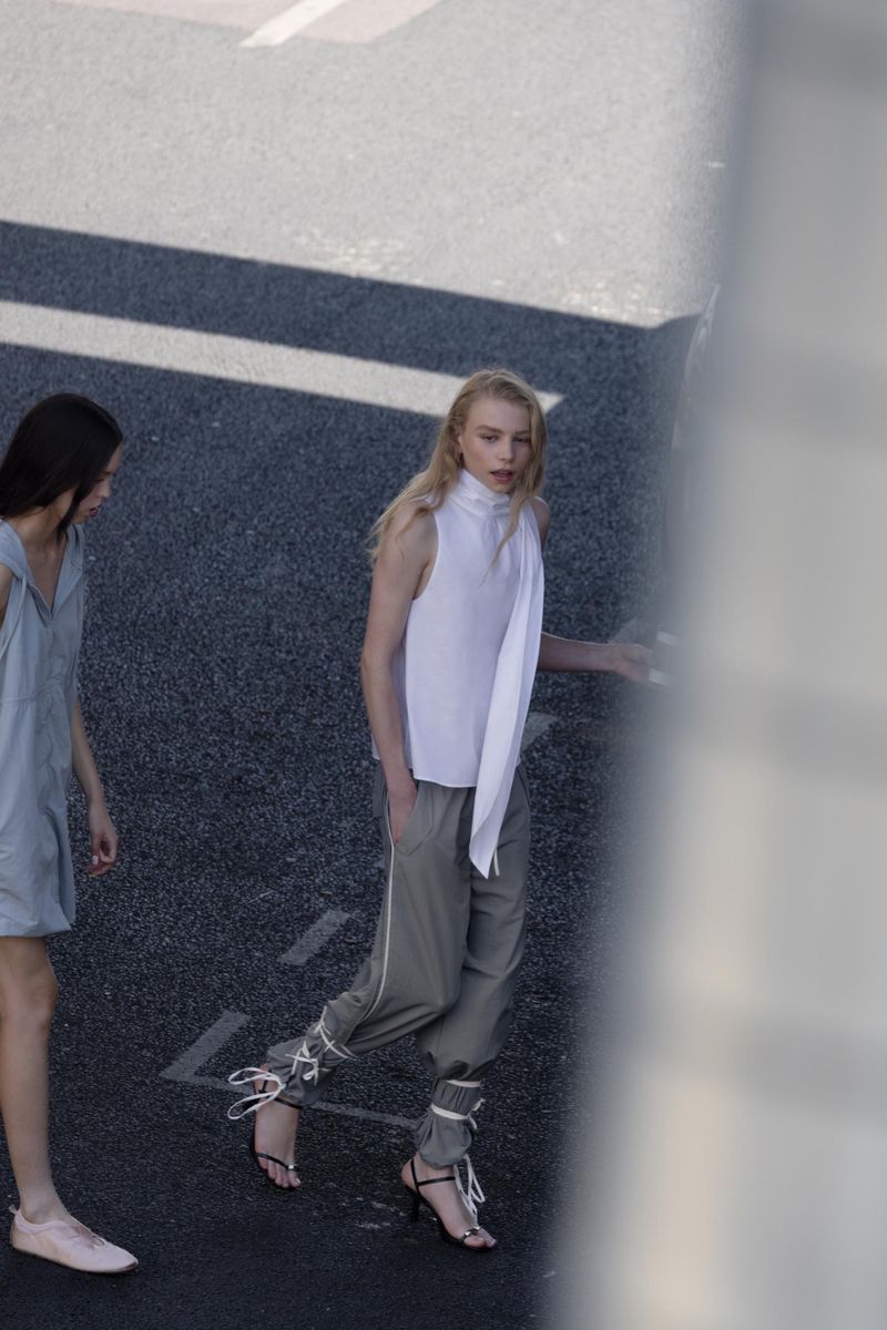 Two women model sleeveless tops and stylish pants on asphalt, showcasing minimalist fashion in an urban setting with natural light.