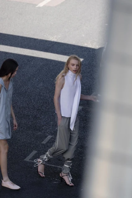 Two women model sleeveless tops and stylish pants on asphalt, showcasing minimalist fashion in an urban setting with natural light.