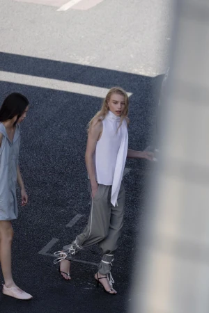 Two women model sleeveless tops and stylish pants on asphalt, showcasing minimalist fashion in an urban setting with natural light.