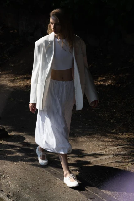 A woman in a white blazer and skirt walks on a stone path, partially lit by sunlight filtering through trees, creating a fashion editorial look.