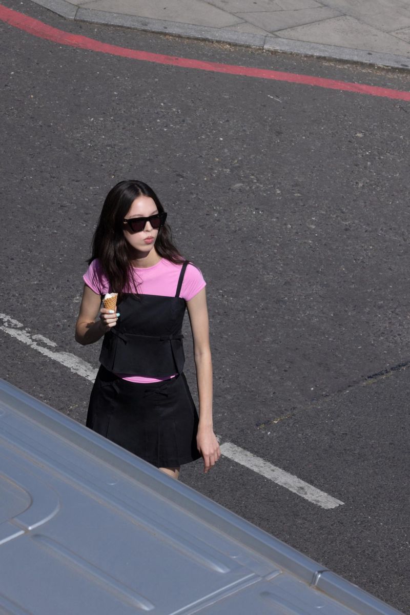 Young woman in sunglasses on city street eating ice cream. She wears a pink top, black skirt, with a silver van in the foreground.