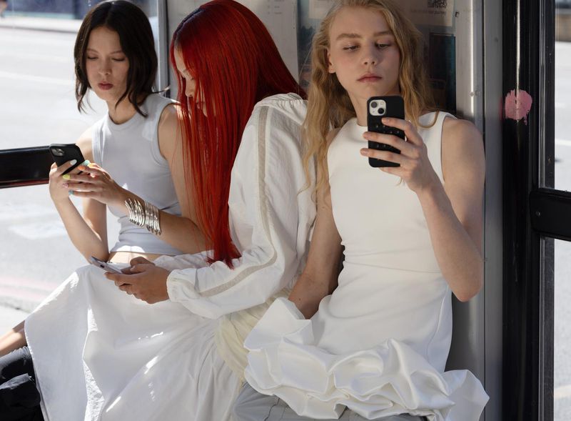 Three young adult women in a phone booth, holding phones and wearing light-colored tops and bottoms. Bright midday light.