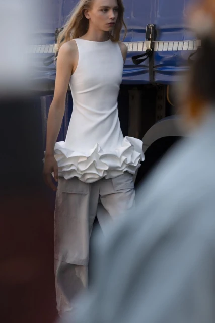Fashion model in white sleeveless dress and light grey wide-leg pants poses outdoors with a blue and white truck in the background.