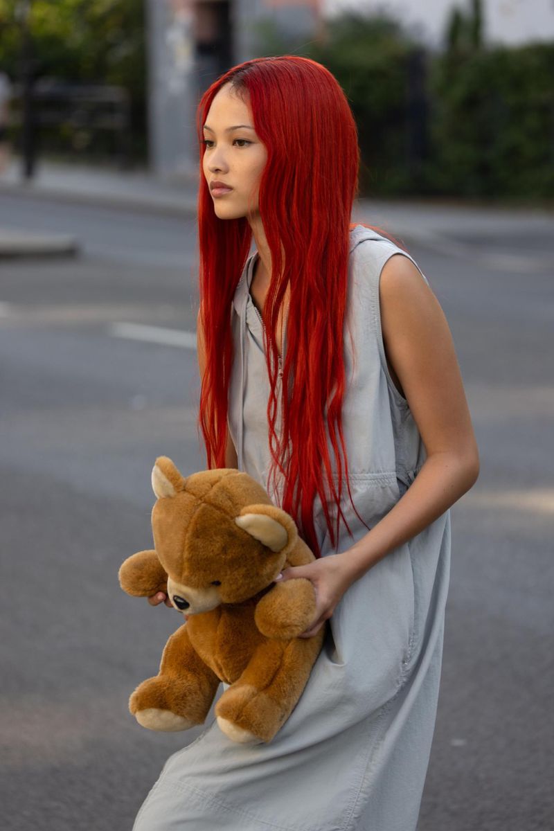 A young adult woman with bright red hair walks down a street holding a teddy bear, wearing a sleeveless, light blue dress.