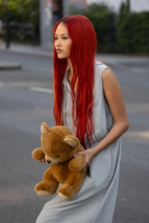 A young adult woman with bright red hair walks down a street holding a teddy bear, wearing a sleeveless, light blue dress.